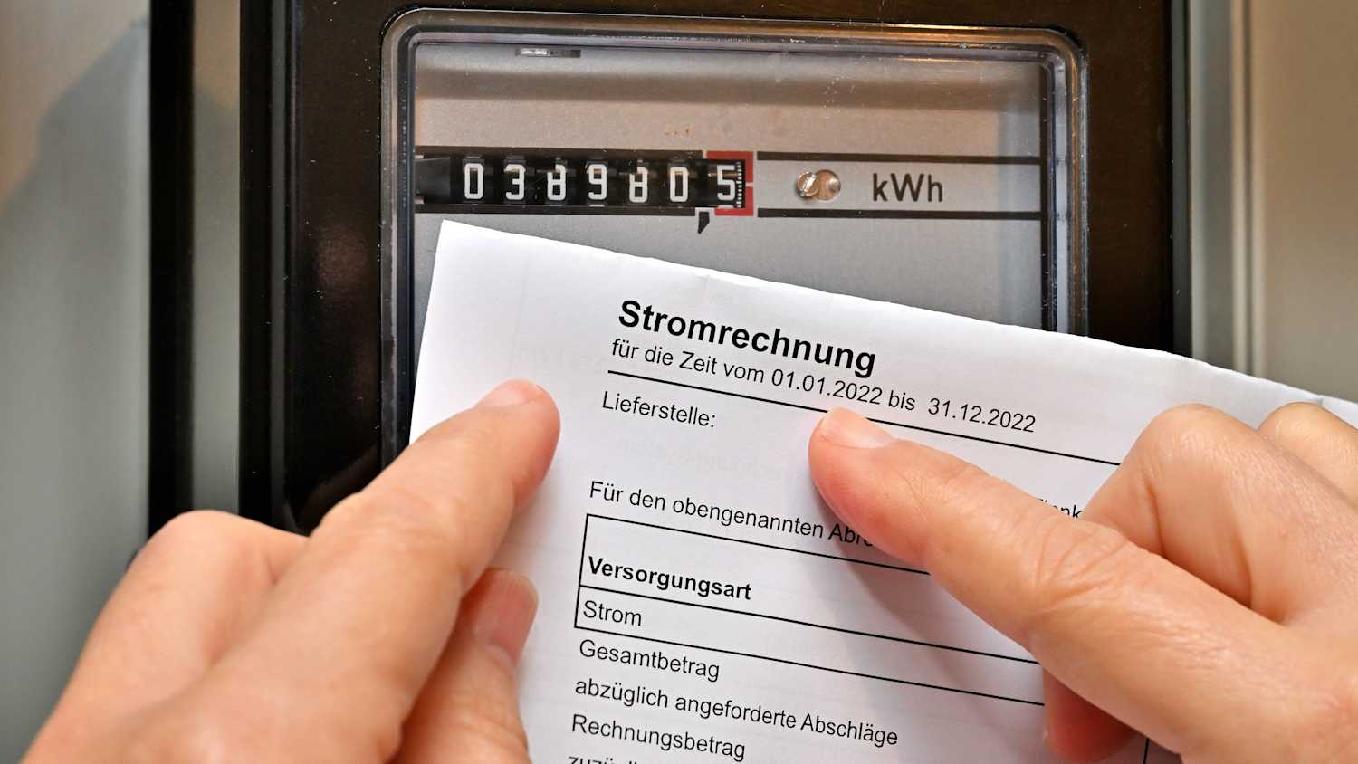 Eine Frau hält die Stromrechnung in der Hand und vergleicht diese mit dem Zählerstand an dem Stromzähler im Haus