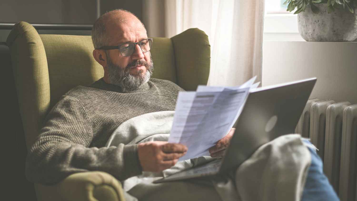 Ein Mann sitzt in einem Ohrensessel neben der Heizung mit einem Laptop auf dem Schoss und Papieren in der Hand