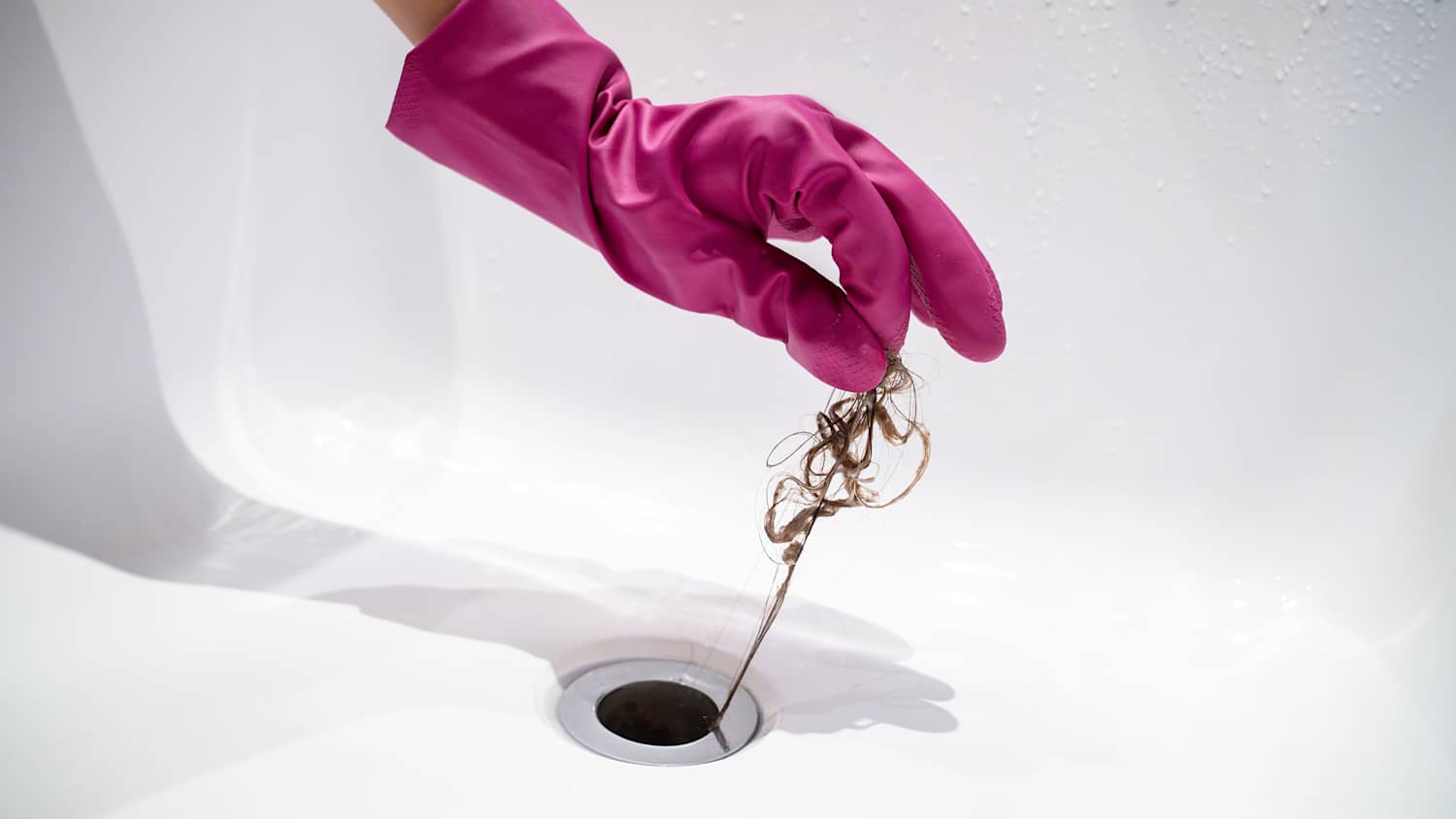 Hand einer Frau mit rosa Gummihandschuh holt Haarbüschel aus dem Abfluss.