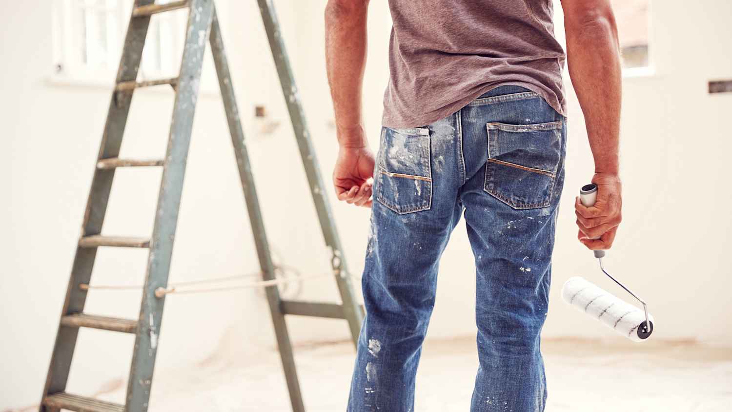 Close Up eines Mannes von hinten, welcher eine farbbemalte Hose trägt und eine Farbrolle in der Hand hält