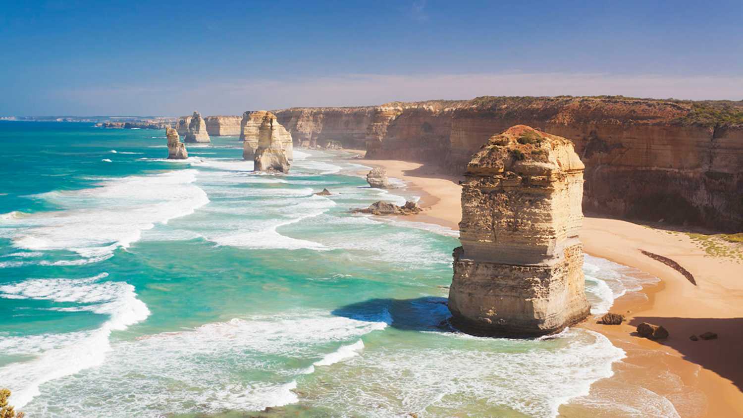 Die Twelve Apostles an der Küste von Victoria, Australien. Mehrere Kalksteinfelsen ragen aus dem Meer, umgeben von Wellen. Die Küste besteht aus steilen Klippen und einem Sandstrand. Der Himmel ist größtenteils klar.