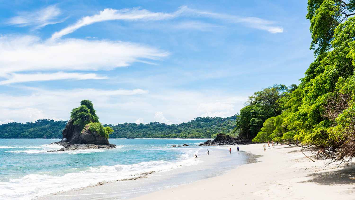Strand in Costa Rica mit weißem Sand und klarem Wasser. Im Hintergrund befindet sich eine bewaldete Felsformation im Meer. Einige Menschen sind am Strand zu sehen. Rechts stehen Bäume. Der Himmel zeigt einige Wolken.