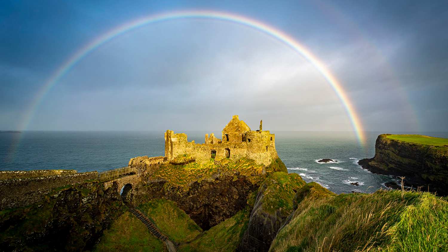 Ruinen von Dunluce Castle auf einer Klippe über dem Meer. Ein Regenbogen spannt sich über den Himmel. Die Umgebung besteht aus Felsen, Gras und Wellen, die gegen die Küste schlagen.