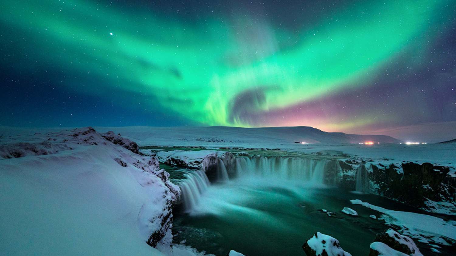 Godafoss-Wasserfall in Island bei Nacht mit Polarlichtern am Himmel. Der Wasserfall ist von schneebedeckten Felsen umgeben. Die Polarlichter erscheinen in Grün- und Lilatönen über dem dunklen Himmel.