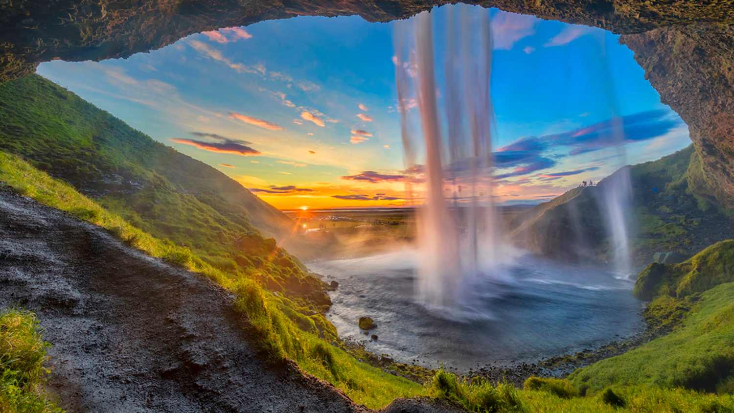 Blick von hinter dem Seljalandsfoss-Wasserfall in Island durch eine Öffnung. Der Wasserfall stürzt in ein Becken, aus dem Nebel aufsteigt. Links führt ein Pfad zur Wasserfallfront, im Hintergrund ist ein Sonnenuntergang mit Wolken am Himmel zu sehen.