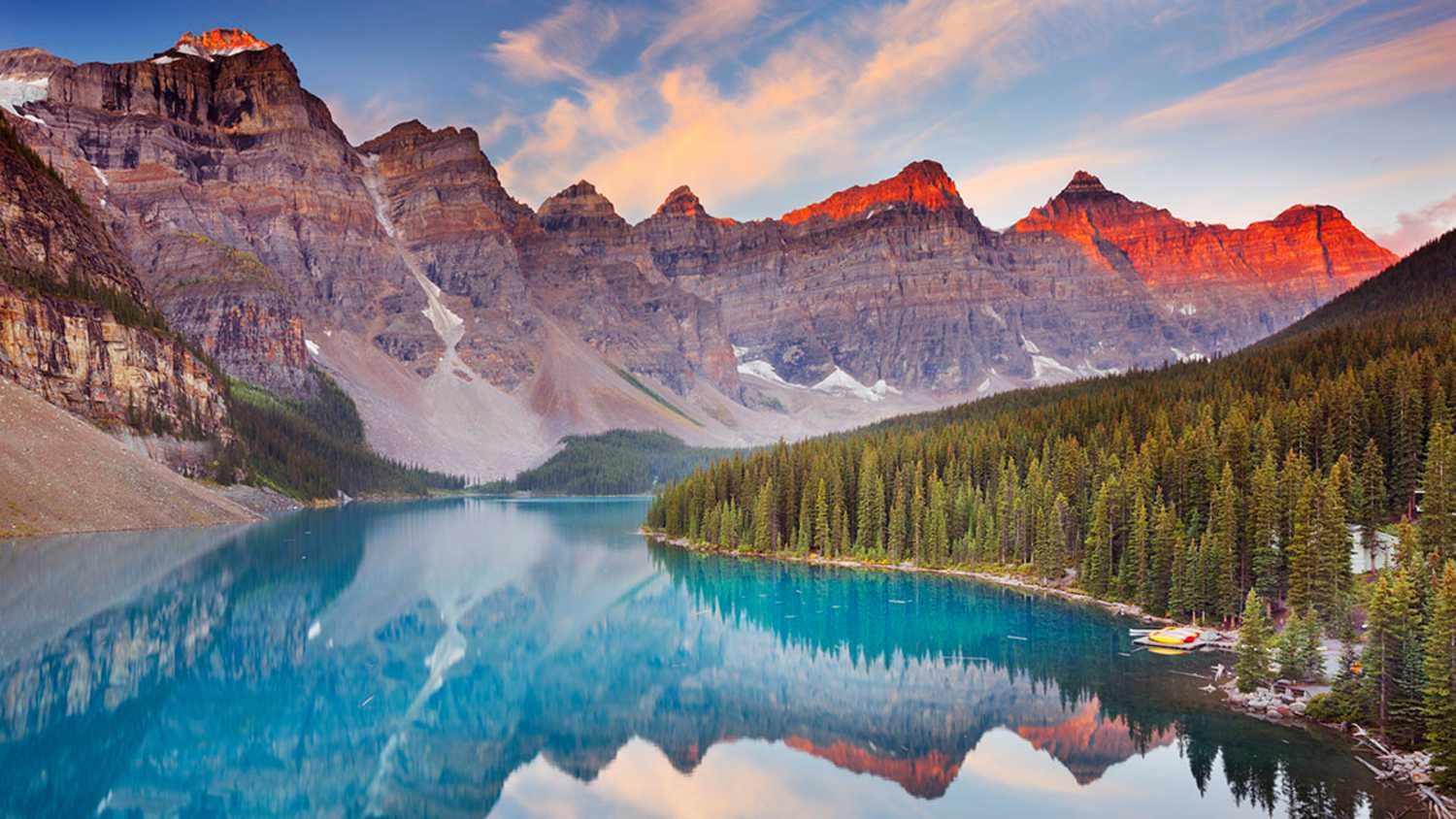 Moraine Lake in Banff National Park, Kanada, umgeben von Bergen des Valley of the Ten Peaks. Die Berge spiegeln sich im klaren Wasser. Am Ufer befinden sich Wälder und Boote. Der Himmel zeigt Wolken und Lichtstimmung bei Sonnenauf- oder -untergang.