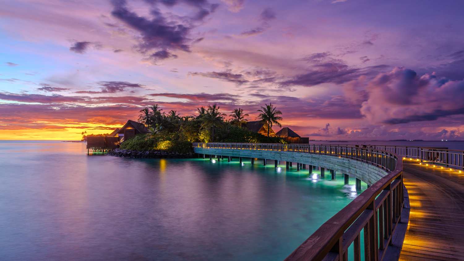 Tropisches Resort auf den Malediven bei Sonnenuntergang mit einem beleuchteten Holzsteg, der zu Wasserbungalows führt, umgeben von grüner Vegetation und einem farbenreichen Himmel, der sich im ruhigen Wasser spiegelt.