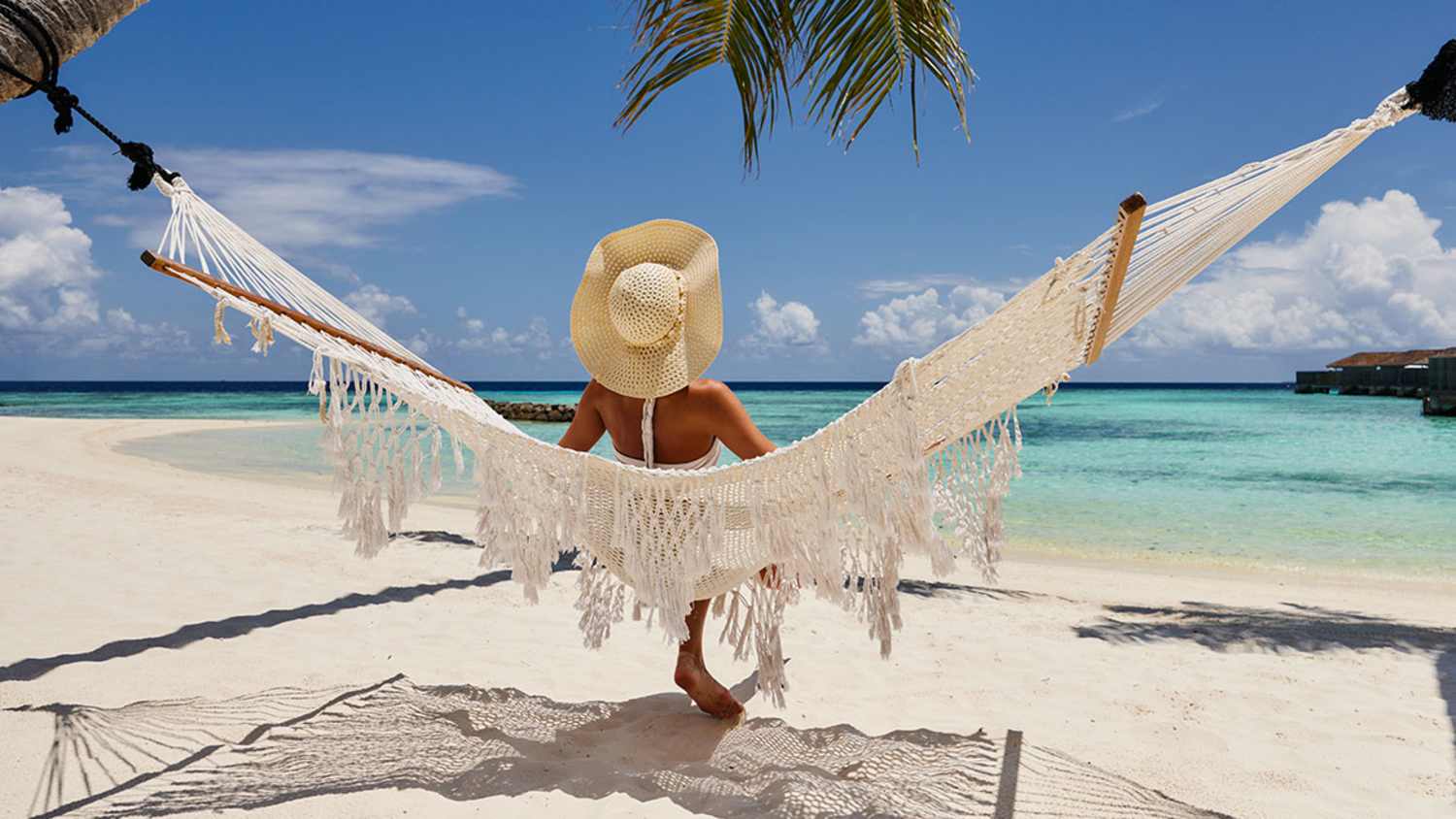 Dame sitzt in einer Hängematte am Strand der Malediven mit Blick auf das Meer. Sie trägt einen Strohhut und ist von Palmen umgeben. Der Himmel ist blau mit einigen Wolken.