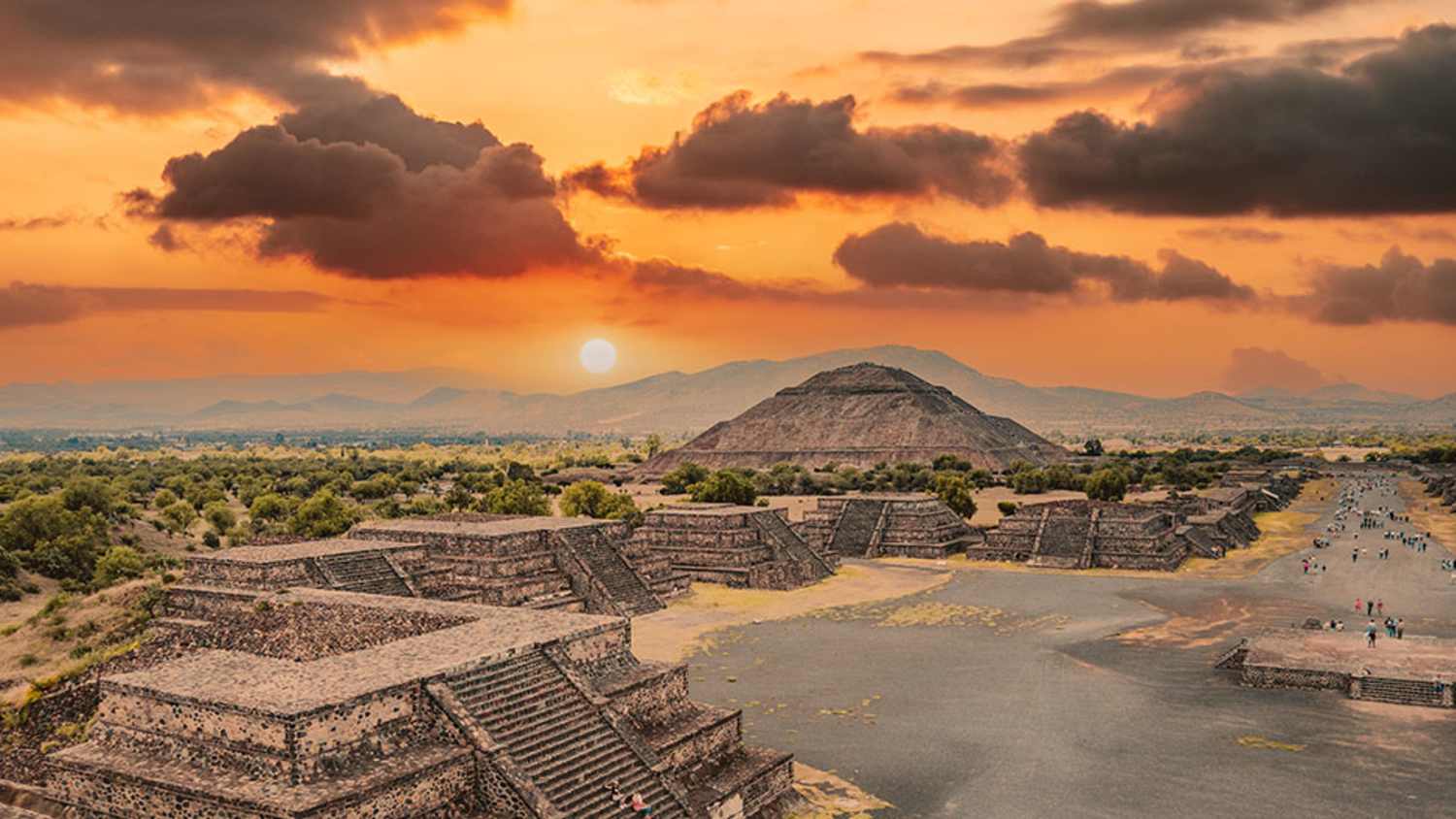 Luftaufnahme der Avenue of the Dead und der Sonnenpyramide in Teotihuacan, nordöstlich von Mexiko-Stadt. Die Aufnahme zeigt einen Sonnenuntergang mit Wolken, kleinere Pyramiden entlang der Straße und die Sonnenpyramide im Hintergrund, umgeben von Grünflächen und Bergen.