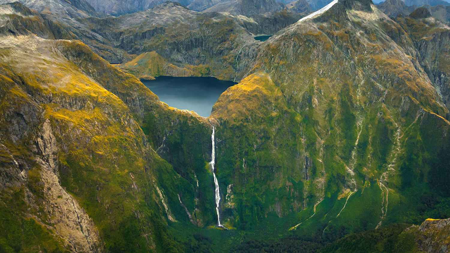 Luftaufnahme von Lake Quill inmitten eines Gebirges, mit dem Wasserfall Sutherland Falls, der vom See in ein darunterliegendes Tal stürzt. Umgeben von felsigem Gelände, Vegetation und schneebedeckten Berggipfeln unter klarem Himmel.