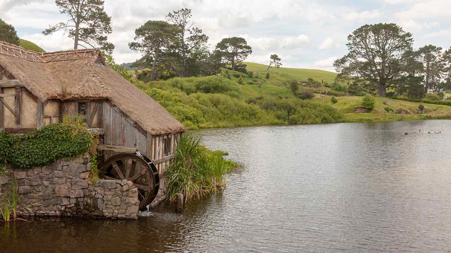 Wassermühle mit Strohdach und Steinfundament am Ufer eines ruhigen Gewässers in Neuseeland. Die Umgebung besteht aus grüner Vegetation, Bäumen und Hügeln unter teils bewölktem Himmel.