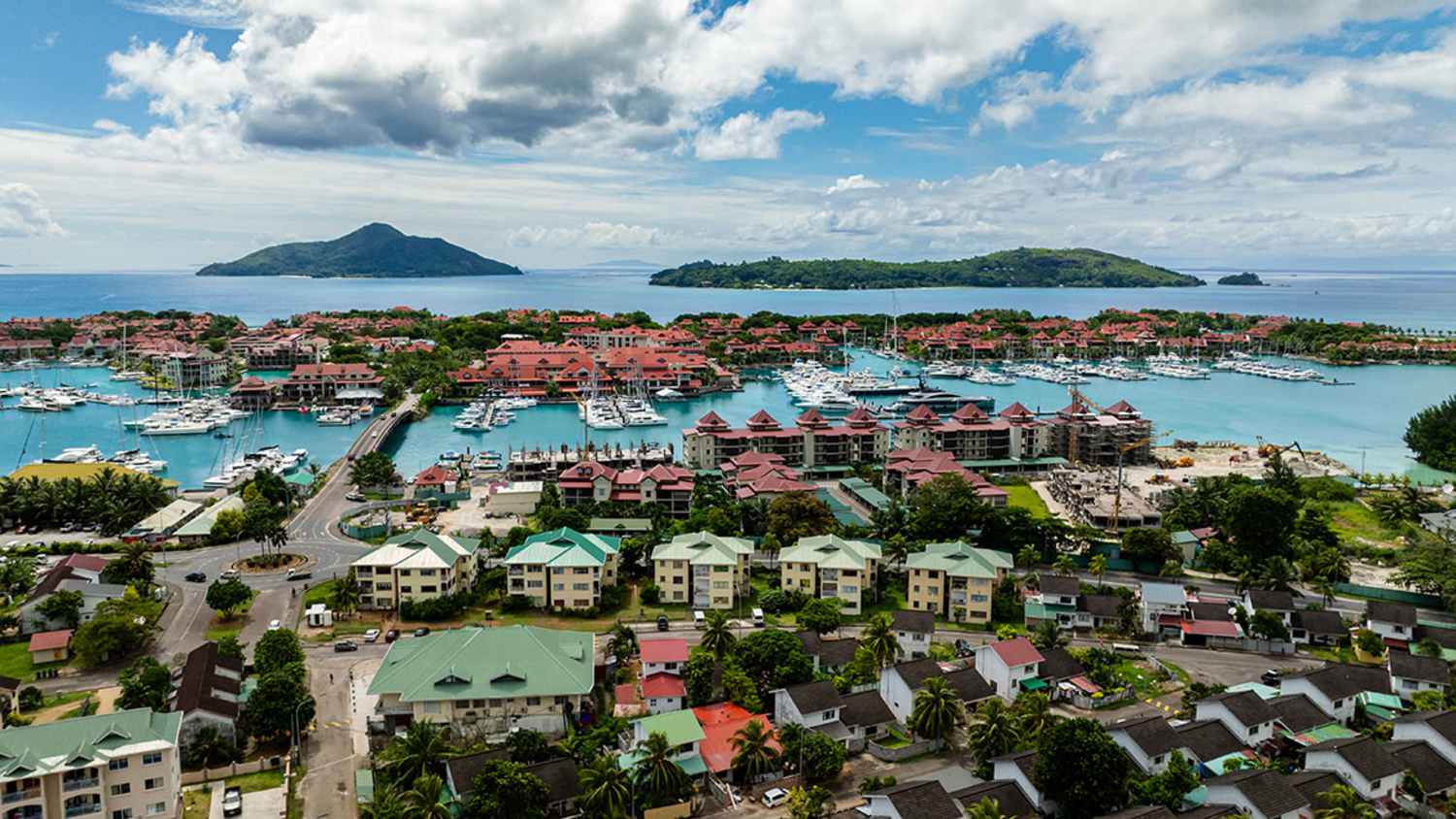 Luftaufnahme von Victoria, der Hauptstadt der Seychellen, mit Wohnhäusern im Vordergrund, einem Yachthafen in der Mitte und grünen Inseln im Indischen Ozean im Hintergrund unter einem teils bewölkten Himmel.