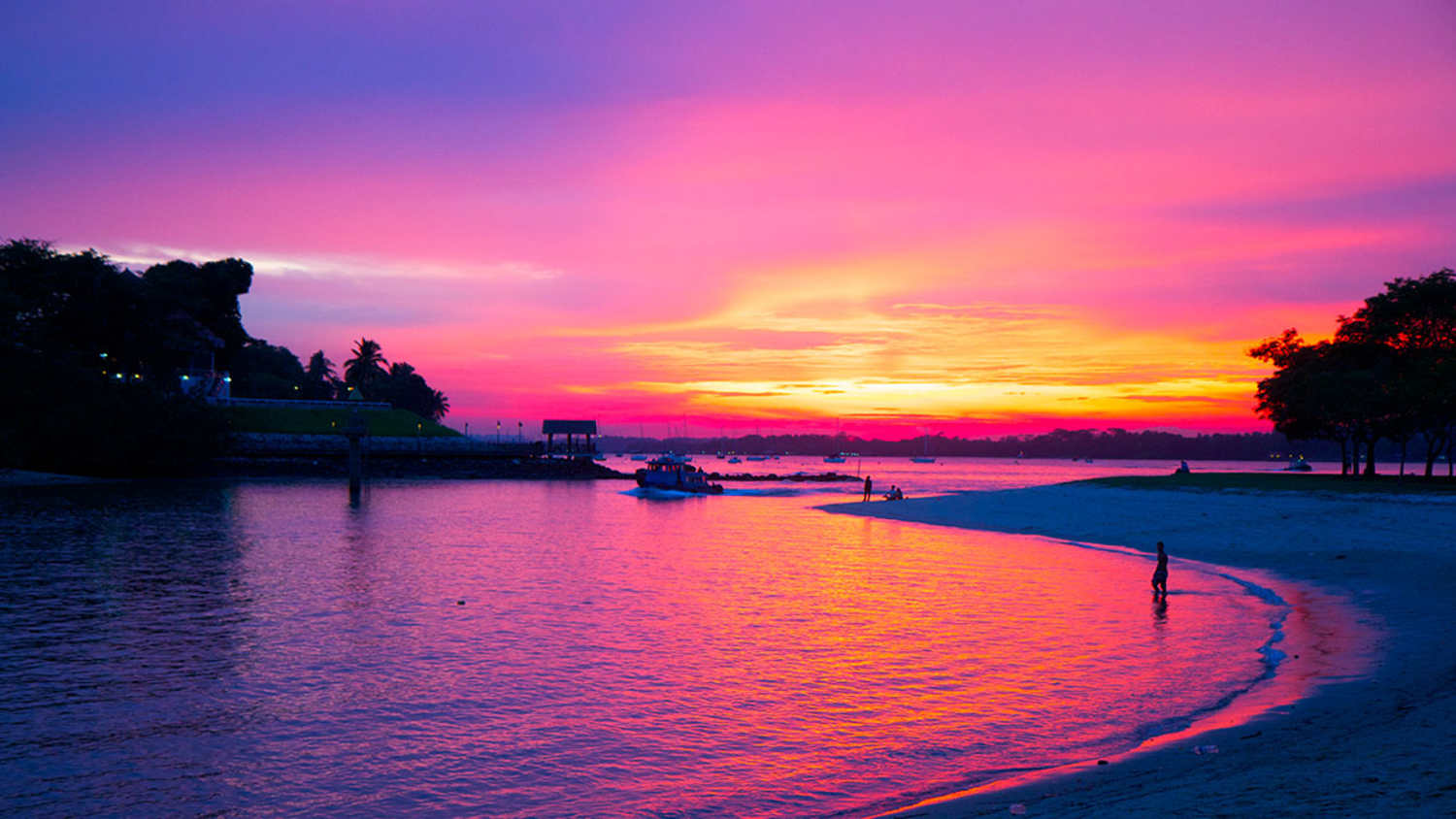 Bumboat kehrt bei Sonnenuntergang vom Pulau Ubin zur Changi Beach in Singapur zurück. Der Himmel zeigt Rosa-, Orange- und Lilatöne. Das Wasser ist ruhig und spiegelt die Farben. Bäume rahmen das Bild, rechts steht eine Person am Ufer. Im Hintergrund ist ein Pier mit Booten und Gebäuden zu sehen.