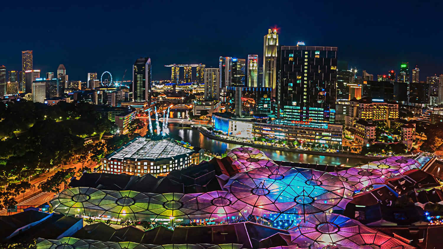 Nachtaufnahme der Skyline von Singapur mit beleuchteten Dachstrukturen von Clarke Quay im Vordergrund, Hochhäusern im Hintergrund und dem Singapore Flyer auf der linken Seite. Die Szene zeigt die lebendige Atmosphäre der Stadt bei Nacht.
