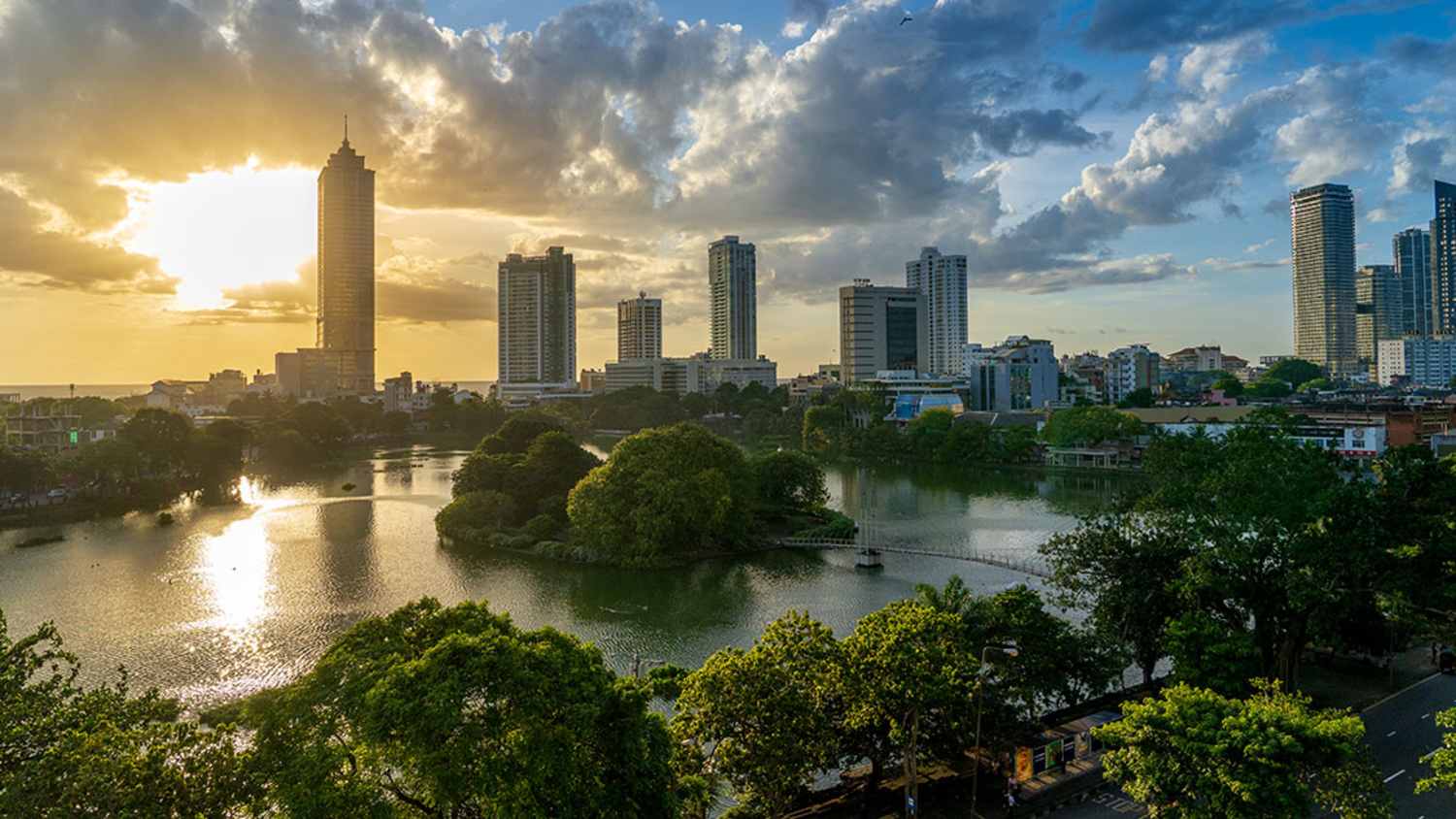 Ein Sonnenuntergang über der Skyline von Colombo, Sri Lanka. Die Sonne geht hinter einem hohen Gebäude unter und wirft ein goldenes Licht auf den Beira-See und die umliegenden Wolkenkratzer. Grüne Bäume umgeben den See und spiegeln das Licht des Sonnenuntergangs wider.