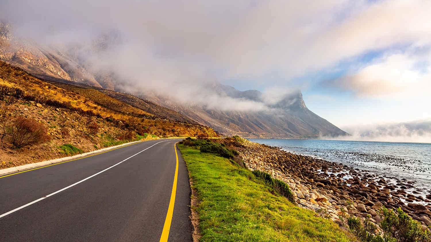 Küstenstraße in Südafrika mit gelber Linie am linken Rand und weißer gestrichelter Mittellinie, entlang einer felsigen Küste mit Blick auf das Meer, grüner Vegetation und nebelverhangenen Bergen im Hintergrund unter teils bewölktem Himmel.
