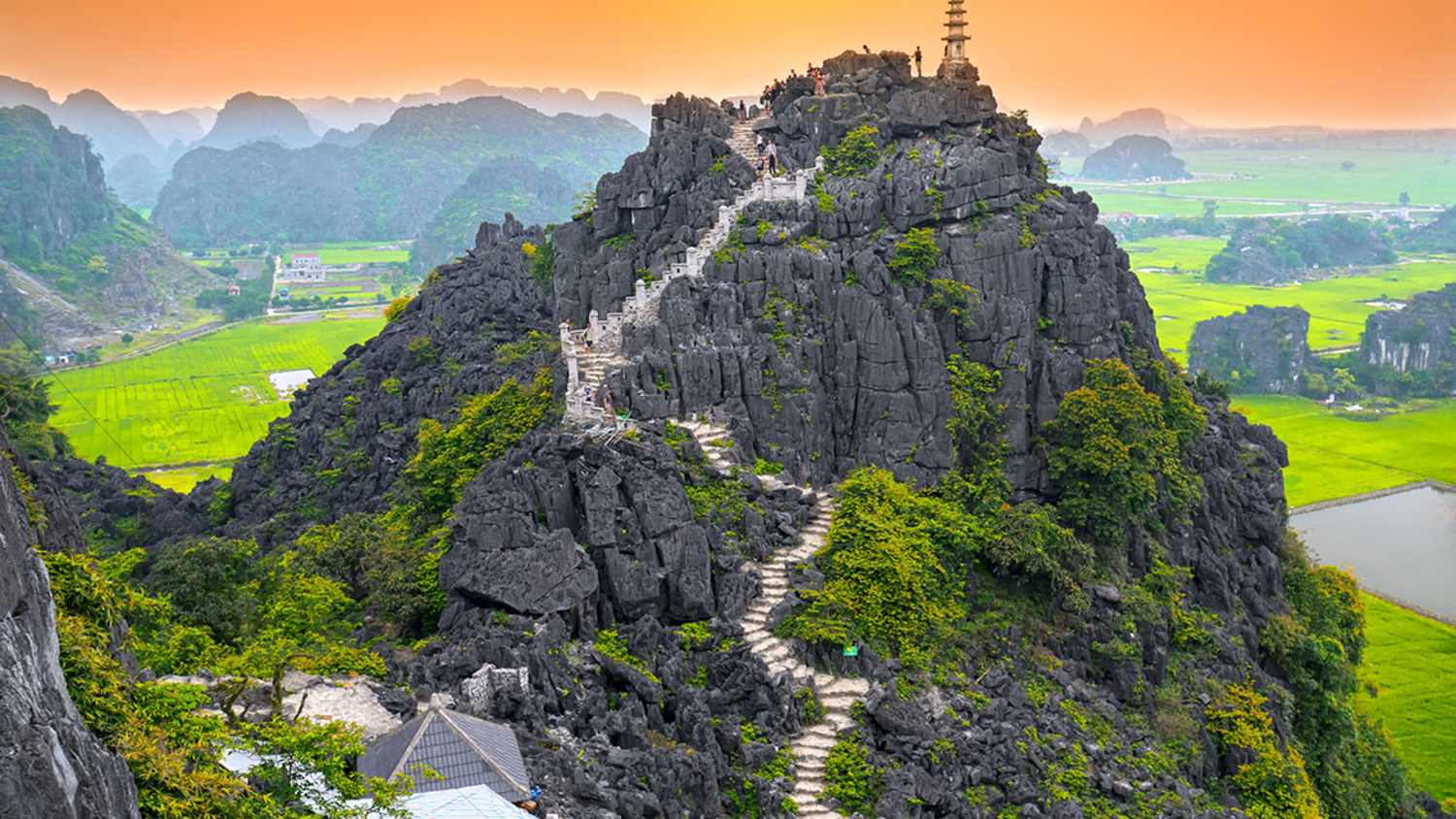 Eine Steintreppe führt den Mua Cave Mountain hinauf zu einer kleinen Pagode auf dem Gipfel. Im Hintergrund färbt die Sonne den Himmel orange. Die Umgebung besteht aus grünen Feldern und entfernten Bergen.
