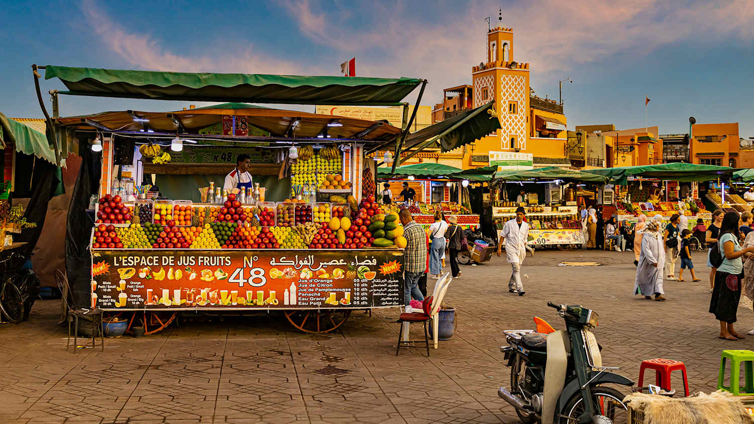 Blick auf einen Markt in Marrakesch mit einem Obststand im Vordergrund, im Hintergrund alte Gebäude und weitere Marktstände