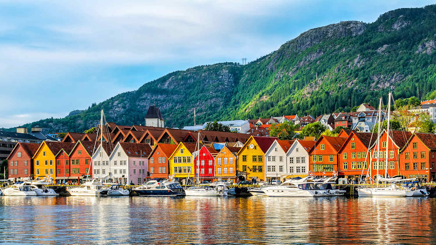 Blick vom Wasser auf eine Reihe bunter alter Häuser in weiß, orange und rot. Im Hintergrund Berge, im Vordergrund Segelboote