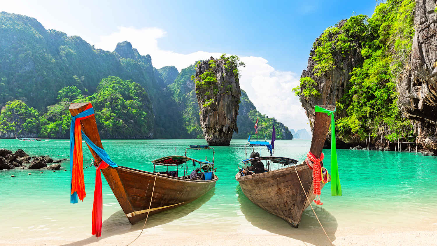 Zwei antike Boote an einem Sandstrand in Phuket vor türkisgrünem Wasser. Die Bucht liegt zwischen hohen Bergen.