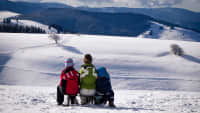 eine Frau und zwei Kinder sitzen auf einem Schlitten und schauen auf die verschneite Winterlandschaft vor ihnen