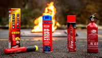 Diverse Handfeuerlöscher für den PKW fotografiert auf Asphalt mit Feuer im Hintergrund