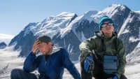 Zwei erschöpfte Wanderer rasten vor verschneiter Bergkulisse