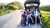 Familie mit Corona Schutzmasken sitz im Kofferraum Ihres Autos auf einer Straße in Frühlingslandschaft