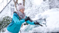 Eine junge Frau Frau mit Bommelmütze schiebt den Schnee von der Windschutzscheibe des verschneiten Autos