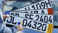 Ein Mann hält ein H-Kennzeichen für historische Fahrzeuge (v. oben), ein Ausfuhrkennzeichen, ein Euro-Kennzeichen und ein Kurzzeitkennzeichen vor ein Auto.