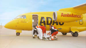 Ein ADAC-Ambulance-Flugzeug steht auf einem Rollfeld. Die Kabinentür steht offen, da ein Patient durch das Rettungspersonal ins Flugzeug gebracht werden soll für einen Krankenrücktransport.