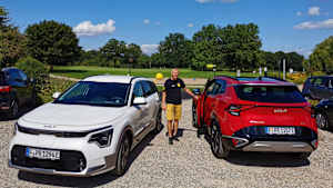 Ein Mann steht zwischen einem roten und einem weißen Kia Sportage auf einem Kiesparkplatz und zeigt wie man die Reichweite auf der Autobahn erweitert.