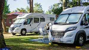 Zwei weiße Wohnmobile stehen auf einem Campingplatz nebeneinander, ein Mann sitzt auf einem Stuhl dazwischen und sieht in seinem Handy nach Wohnmobile zu kaufen