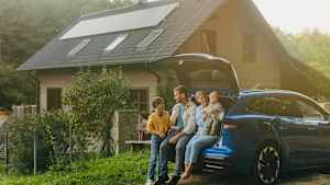 Eine Familie, die sich an einem sonnigen Tag vor ihrem E-Auto und Haus mit PV-Anlage aufhalten.s mit PV-Anlage sitzt.