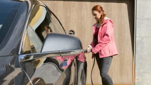 Eine Frau in einer pinken Jacke lädt ein schwarzes Elektroauto vor einer Holzgarage durch eine mobile Ladestation.