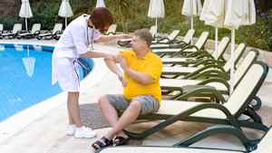 Eine Aerztin behandelt einen maennlichen Reisenden, der auf einer Liege am Pool eines Hotels sitzt. Im Hintergrund stehen weitere Liegen und Sonnenschirme.