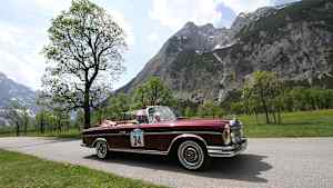 Zwei Männer fahren in einem roten Oldtimer eine Straße neben den Bergen entlang und müssen sich keine Gedanken über eine Oldtimerversicherung machen
