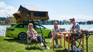Eine Gruppe junger Leute sitzen an einem Campingtisch vor einem aufgebauten Dachzelt an einem See