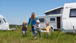 Kinder laufen mit einer Frau über eine Wiese vor einem Karavan.