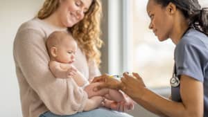 Eine Mutter hat ihr Baby auf dem Schoss wärend es eine Sprizte von einer Ärztin bekommt