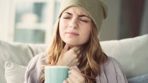 Eine Frau mit Mütze und Tasse langt sich an ihren schmerzenden Hals