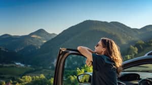 Eine Frau steht an ihrer Autotür gelehnt im Sommer mit Ausblick auf die Berge