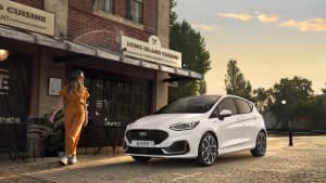 Eine Frau blickt auf den neuen weißen Ford Fiesta ST Line in Weiss, der am Straßenrand parkt