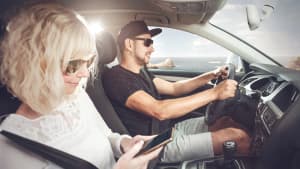 Ein Mann und eine Frau sitzen in einem Auto, der Mann fährt, die Frau ist am Handy