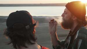 Zwei Freunde rauchen Joint mit Gras am Meeresufer bei Sonnenuntergang