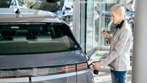 Frau steht in der Nähe von Elektroautos mit Fernbedienung in der Hand im Autohaus. Frau mit Ladekabel für Elektroauto, glückliches Warten auf Elektroauto zum Aufladen.