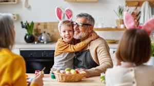 Glückliche Großeltern, Eltern und Kinder aus mehreren Generationen bereiten sich zu Hause gemeinsam auf die Osterferien vor, Großvater und Enkel in Hasenohren umarmen sich, während sie in der Küche Eier bemalen