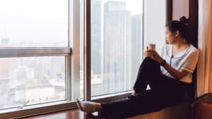 Junge Frau sitzt am Fenster mit einem Becher in der Hand am Fenster und schaut nach Draussen