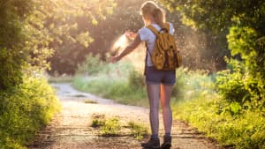 Frau auf Wanderweg im Wald, in der Abendsonne besprüht Ihre Arme mit Mückenschutz-Spray