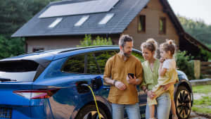Familie mit kleinem Mädchen steht vor ihrem Haus mit Sonnenkollektoren auf dem Dach und hat ein Elektroauto.